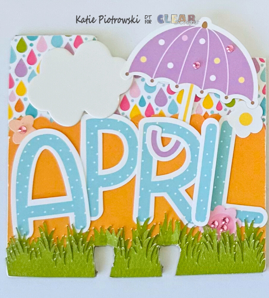 A colorful craft card displaying the word 'APRIL' in bold letters, adorned with an umbrella, cloud, and decorative elements like grass and flowers.
